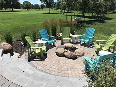 Hardscaped patio with brick stones and fire pit with green and blue chairs completed landscape job